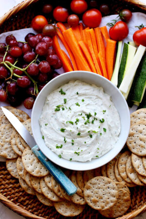 Wicker tray with bowl of whipped goat cheese, crackers, grapes and veggies and a blue handled knife.