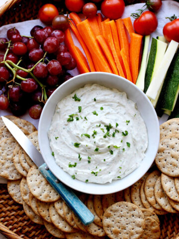 Wicker tray with bowl of whipped goat cheese, crackers, grapes and veggies and a blue handled knife.