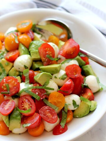 Tomato mozzarella avocado salad in a white bowl with a silver spoon.