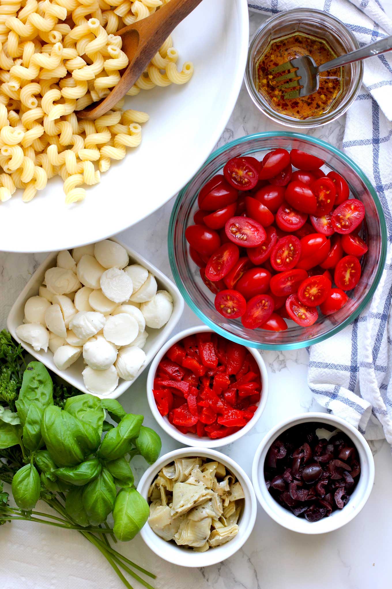 Ingredients to make pasta salad in individual ceramic bowls with fresh herbs and a blue and white napkin.