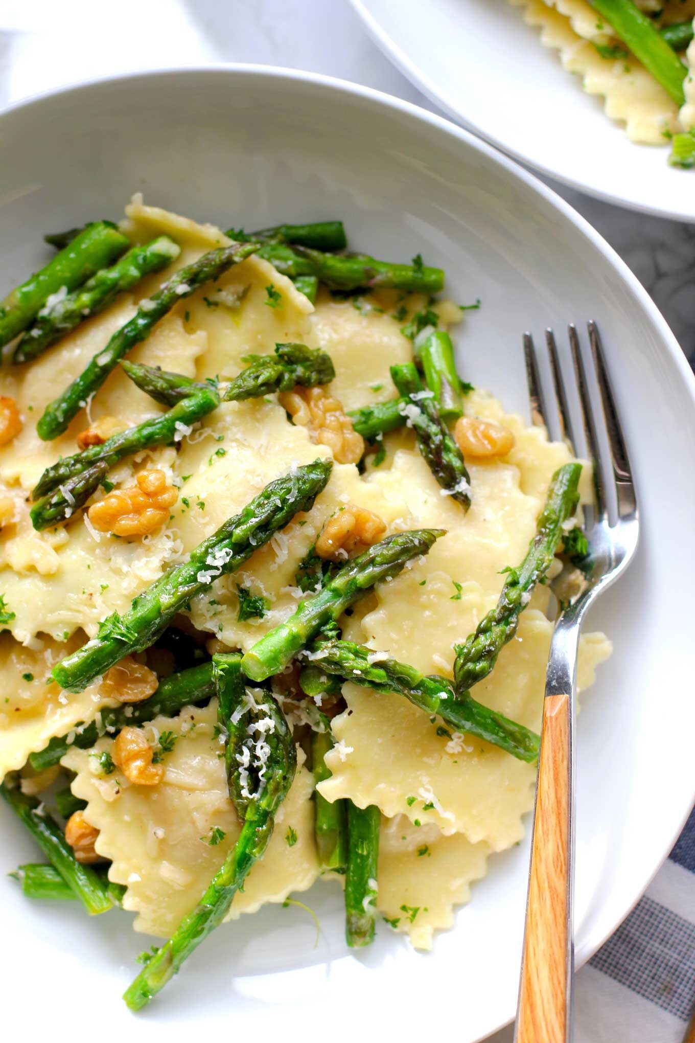 Close up of ravioli with asparagus and walnuts in a white bowl with a fork.