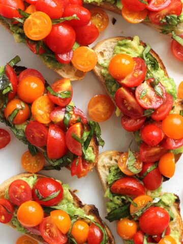 Close up of avocado toast topped with orange and red cherry tomatoes on a white dish.